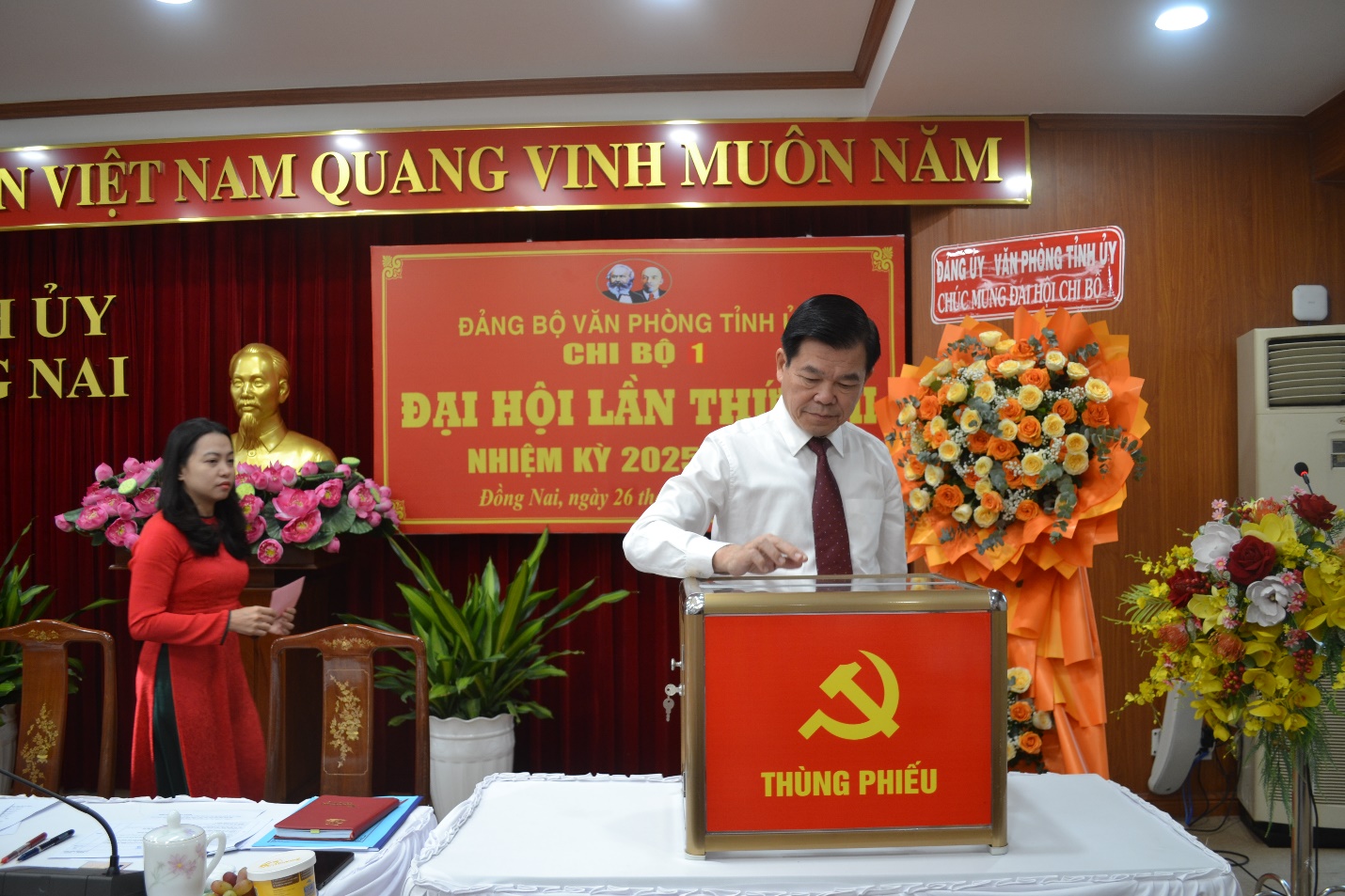A person standing at a podium with a red sign and flowersDescription automatically generated